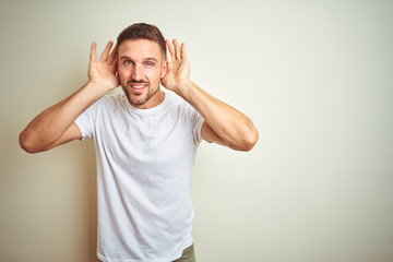 Young handsome man wearing casual white t-shirt over isolated background Trying to hear both hands on ear gesture, curious for gossip. Hearing problem, deaf