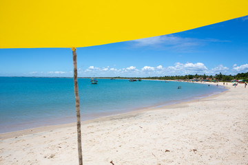 Fototapeta na wymiar Beautiful beach view with a yellow tent on sunny summer day and sea and blue sky in the background. Concept of vacations, peace and relaxation. Ponta do Corumbau, Bahia, Brazil.