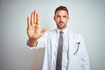 Young handsome doctor man wearing white profressional coat over isolated background doing stop sing with palm of the hand. Warning expression with negative and serious gesture on the face.