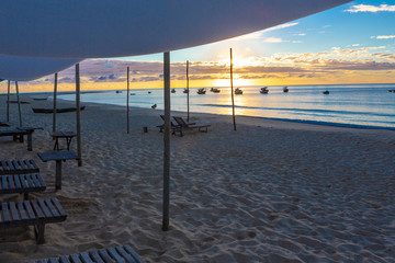 Beach with wooden chairs, sun loungers and tent with boats and sunset in the background.Concept of vacations, peace and relaxation. Ponta do Corumbau, Bahia, Brazil