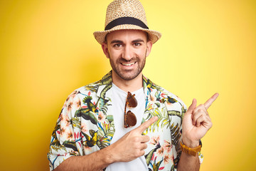 Young man on vacation wearing hawaiian flowers shirt and summer hat over yellow background smiling...