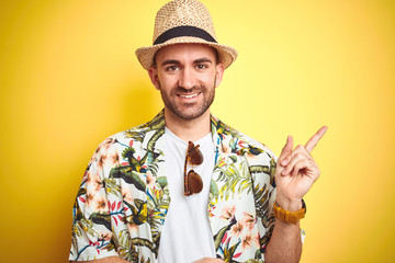 Young man on vacation wearing hawaiian flowers shirt and summer hat over yellow background with a big smile on face, pointing with hand and finger to the side looking at the camera.