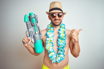 Young man wearing summer hat and hawaiian lei flowers holding water gun over isolated background...