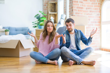 Young beautiful couple moving to a new house sitting on the floor clueless and confused expression...