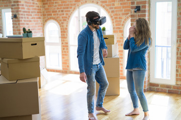 Young couple moving to a new house having fun playing with virtual reality glasses around cardboard boxes