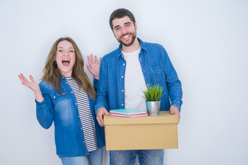 Young couple holding cardboard box moving to new house over white isolated background very happy and excited, winner expression celebrating victory screaming with big smile and raised hands