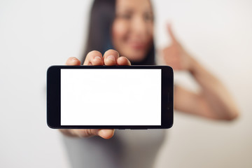 A young girl woman is holding a smartphone with a blank white screen horizontally in front of her and smiles