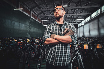 Pensive man in protective glasses and checkered shirt is posing at his own warehouse full of bicycles.