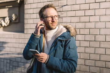 Stylish man with beard and glasses dressed in blue jacket inserts wireless headphones into ears. Modern technologies concept. Male listen audio message or music, in the hands he holds mobile the phone