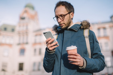 Attractive male with beard, glasses and in blue jacket chatting or surfing in web. Hipsters in urban environment. Pleasant man drink coffee to keep warm. Selective focus on phone. Internet addiction.