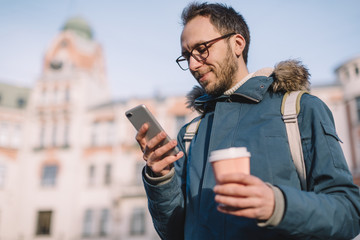 Handsome adult male with beard, glasses and in blue jacket looking into smartphone and chatting with girlfriend. Internet dating. Charming man drink coffee to keep warm. Concept of pleasant emotions