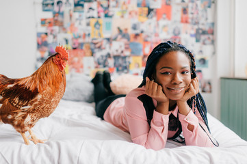 African american girl lying on white bed with chicken on decorative wall background.