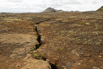 Barren steppe-like landscape in shades of brown and ocher, through the surface runs a dark rift,...