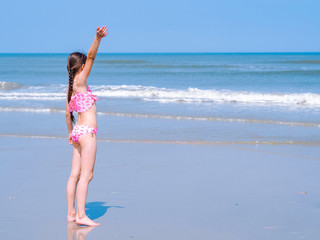 Fototapeta premium Beautiful teen girl with long hair wearing in pink swimsuit standing on the beach and looking into the distance and wave hands at sea. Enjoying a lovely summer day. Summer travel and vacation concept