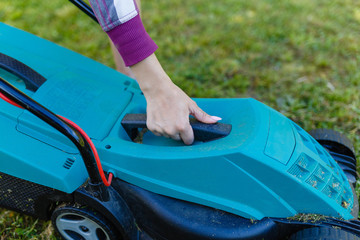 Women's hands on the handle are a lawn mower. The girl carries the lawnmower on the mowed grass and begins to mow the lawn.