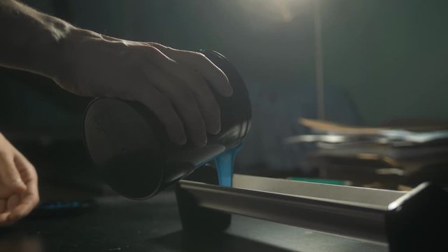 Young caucasian man pouring container with blue color in workshop in industrial print studio. Craftsman print on t shirt silkscreen.