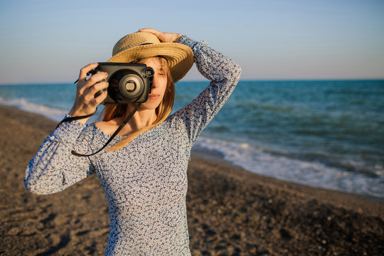 Young attractive girl with long blond hair, wearing a hat, takes pictures at the beach. Holding photocamera in one hand. Blue waves on the background.