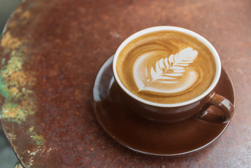 Top and close up view of a brown ceramic cup of cappuccino on top with beautiful delicate latte art with leaf pattern, and background of red rough rusty metal table. Vintage and industrial tone.