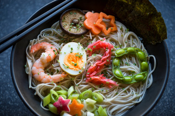 Bowl of Japanese buckwheat ramen noodle soup topped with the number 2020 spelled out of shrimp, boiled egg, pickled ginger, and edamame beans