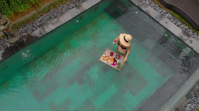 Aerial shot of a young woman tourist has her own personal breakfast on a floating table in a private swimming pool