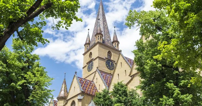 Timelapse of Lutheran Cathedral of Saint Mary in Sibiu, Romania - zoom in to tower clock. Fairytale looking building framed by green trees, with fast white puffy clouds on blue sky, on a sunny day.