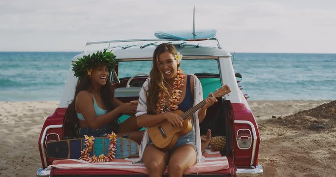 Two beautiful young women singing and playing ukulele at the beach tailgating on a vintage car, hawaiian island surf concept