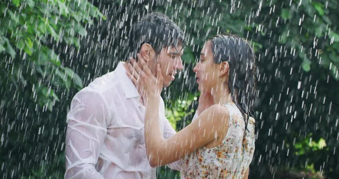 Slow motion of young carefree couple in love are hugging and kissing under the rain on a background of green trees.
