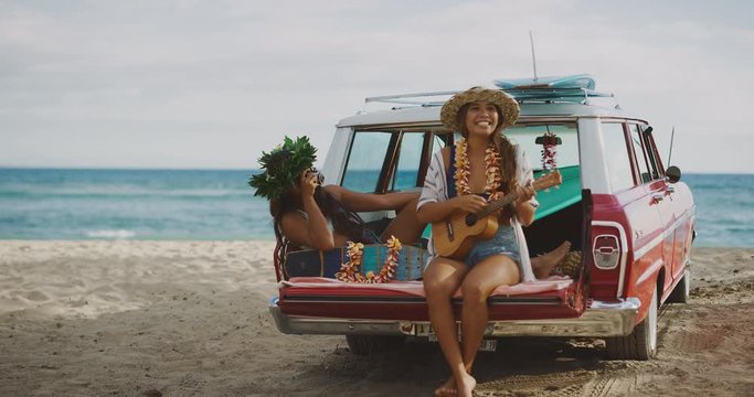 Two beautiful young women singing and playing ukulele at the beach tailgating on a vintage car, hawaiian island surf concept
