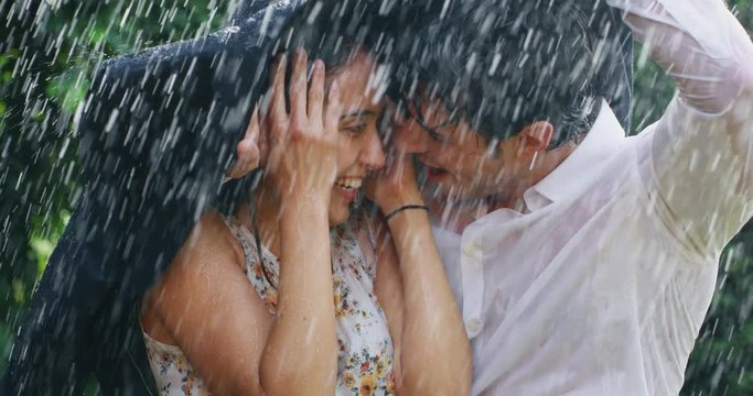 Slow motion of young carefree couple in love are covering with his jacket for not to get wet and having fun under the rain on a background of green trees.