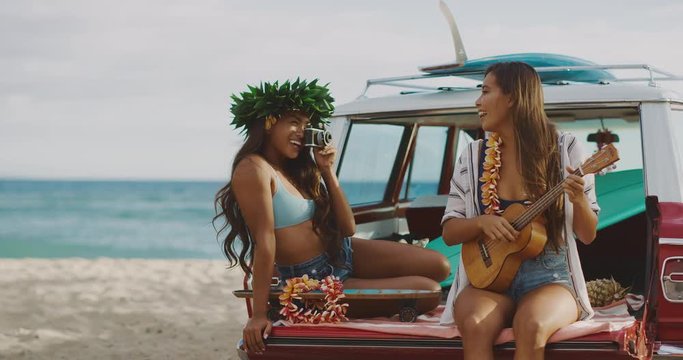 Two beautiful young women smiling taking pictures and playing ukulele at the beach tailgating on a vintage car, hawaiian island surf concept, summer fun at the beach