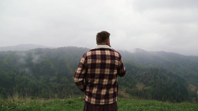 man kilt with a mustache and beard is standing on top green mountain on the grass, wearing a plaid shirt, looks around and smokes. it is snowing from the sky. fog around caucasian