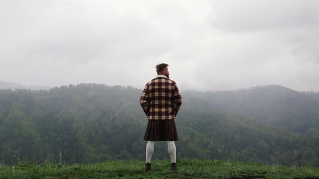 man kilt with mustache beard standing on the top Green Mountain grass, dressed plaid shirt, white socks brown shoes, looks around. it is snowing from the sky. fog around.Caucasian