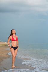 slim brunette in a bikini stands on a sandy beach whit closed eyes