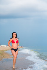 a slender brunette in a bikini stands on a sandy beach near the surf whit hans on waist