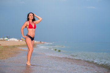 Fototapeta premium a slender brunette stands in red bikini on a sandy beach near the surf