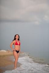 a slender brunette in a bikini stands on a sandy beach near the surf whit closed eyes and hands on her waist