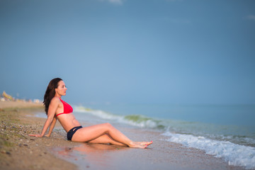 slender brunette girl with long hair in a red bikini sits on sandy beach 