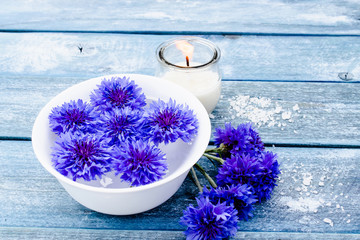 blue cornflowers lie in water in a white bowl near a burning candle against the background of blue old boards.