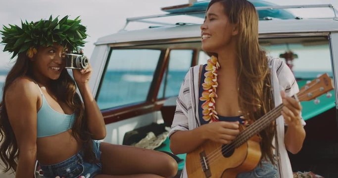 Two beautiful young women smiling taking pictures and playing ukulele at the beach tailgating on a vintage car, hawaiian island surf concept, summer fun at the beach