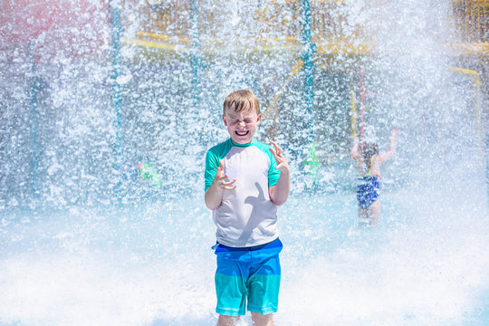 Young boy getting soaking wet while at an outdoor water park. Lots of water splashing water behind the boy. He is smiling and anticipating getting wet and drenched