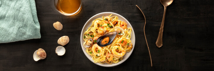 Seafood pasta panorama. Tagliolini with mussels, shrimps, clams and squids, with a glass of white wine, shot from the top on a black background with a place for text