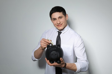 Young businessman putting money into piggy bank on light background