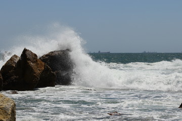Big storm waves hitting the coastal rocks of the sea. Sunny windy day, blue sky. Splashes and foam. Storm landscape.