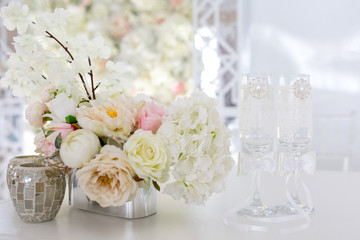 Festive concept of white wedding. Flower arrangement of artificial bouquet on the table. Glasses for the bride and groom.