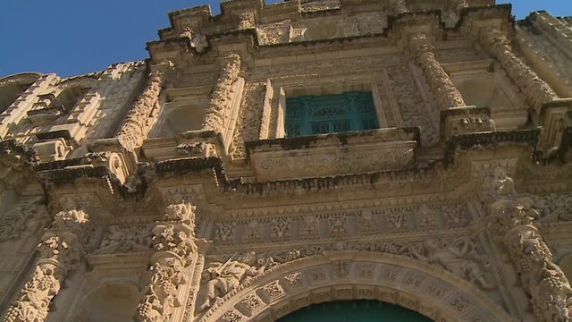 A panning low angle shot of an old building in focus with the sun shining however so graciously through the blue and cloudy sky