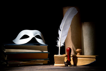 Drama or theater and literature concept. Old inkstand with feather near scrolls with mask on books against black background. Dramatic light - obrazy, fototapety, plakaty