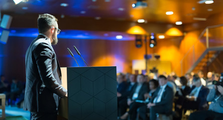 Speaker giving a talk on corporate business conference. Unrecognizable people in audience at...