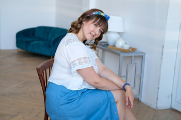 Fototapeta na wymiar A young plus size woman sits on a chair in a spacious white room. A girl in a long denim skirt, sandals with high heels, a scarf on her head. Bright boho style.