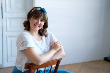 A young plus size woman sits on a chair in a spacious white room. Smile on his face, flirtatious look. Happiness and beauty in any weight. A girl in a long skirt, sandals with high heels