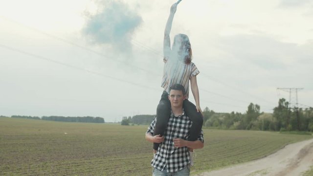 Handsome male walking on country road holding smiling female with color smoke bomb on shoulders. Happy woman sitting on man's shoulders setting arms in air imagining flight over countryside.